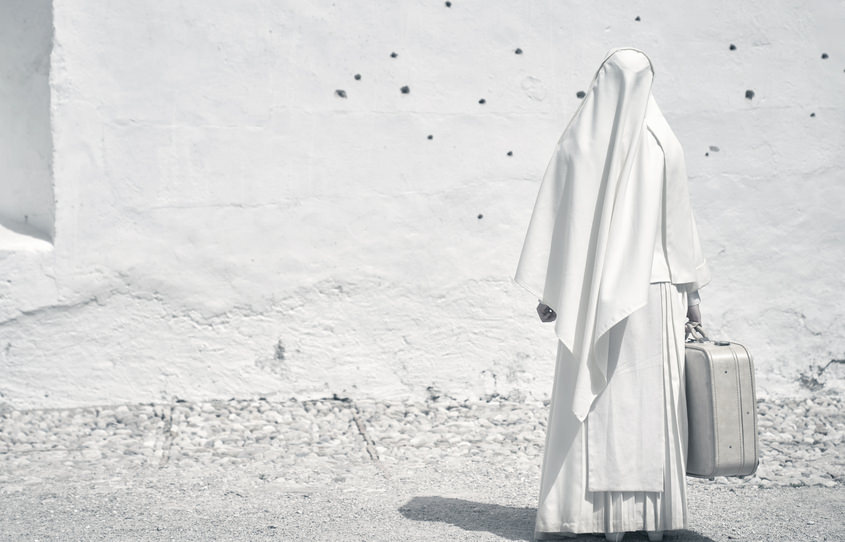 Una monja vestida de hábitos blancos permanece de pie, de espaldas, sujetando una maleta y mirando al horizonte. La imagen entera está en distintos tonos de blancos.